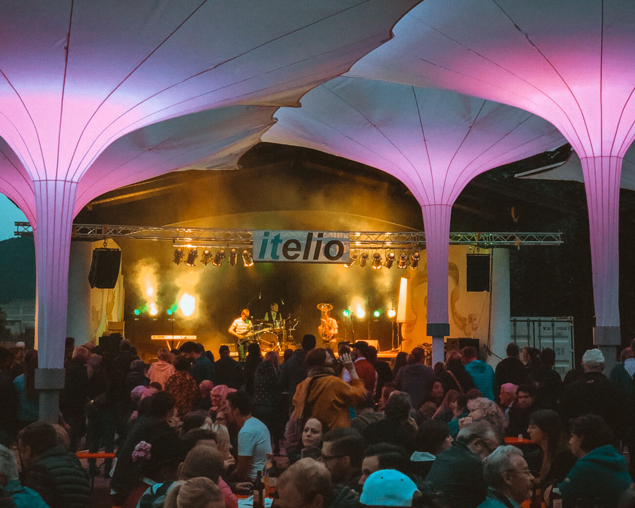 Bühne mit Band und Lcihter, Gäste im Vordergrund, Große Sonnenschirme lila beleuchtet